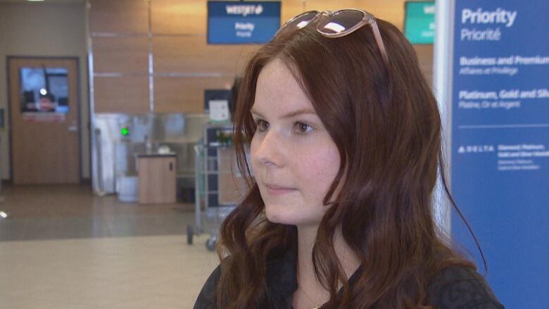 A woman with dark brown hair, with sunglasses on top of her head, is standing in an airport lobby.