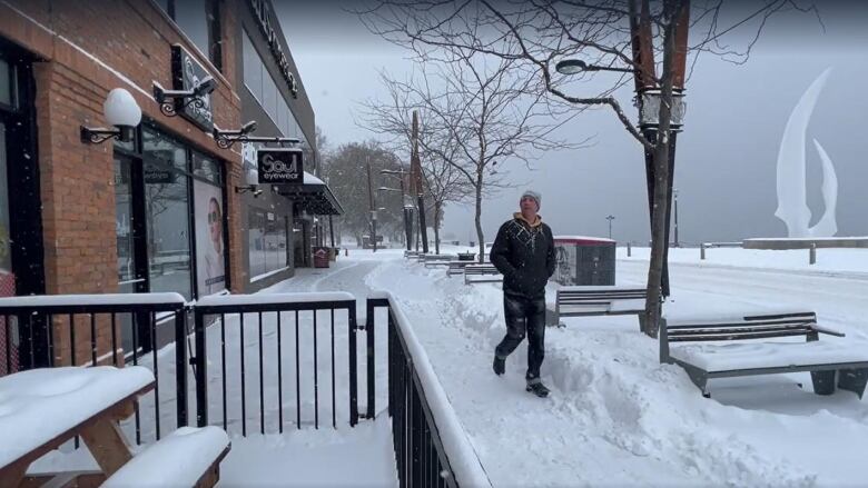 A man walks along a snowy sidewalk next to a snow-covered road with shops on one side and a statue behind him on the other side.