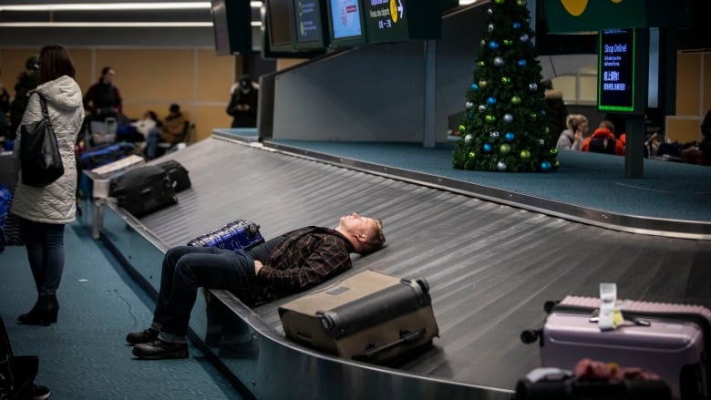 A man with a suitcase beside his head lies down and closes his eyes on a baggage carousel with a Christmas tree in the background and passengers milling around.