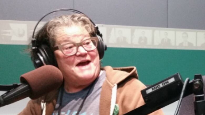 A woman wearing a brown hooded sweater and glasses sits in front of a microphone.