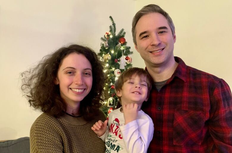 A woman and man smile with their toddler son in front of a Christmas tree.