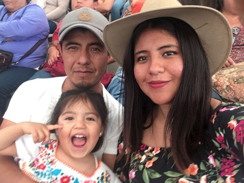 Three family members smile, posing for the camera at an event in Mexico. Salomon Lzaro holds his young granddaughter, who wears a colourful flowered Mexican blouse.