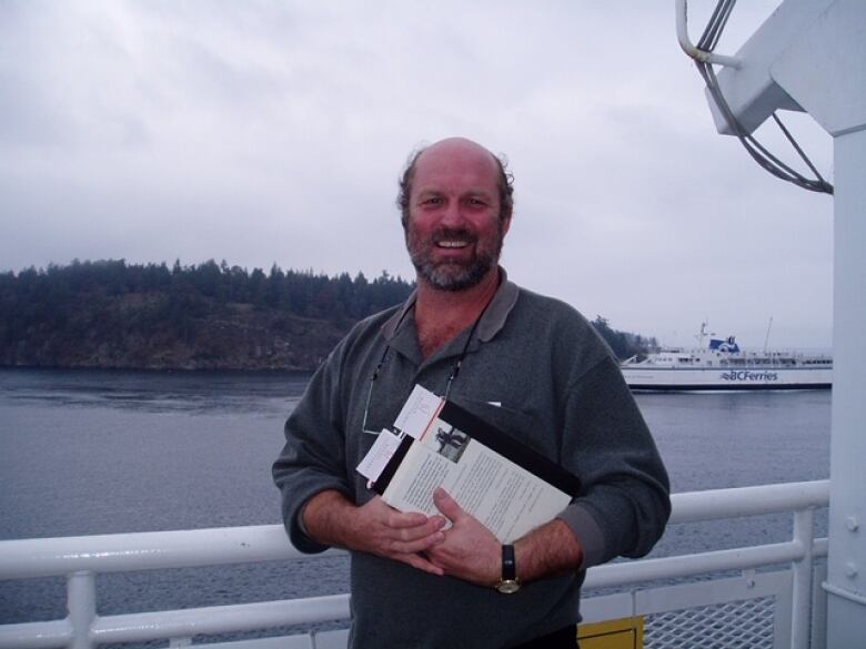 A man stands beside a railing with a clipboard in his hands. He's outdoors beside the water - there's a boat passing by behind him. The man is wearing a green button-up shirt and is smiling.