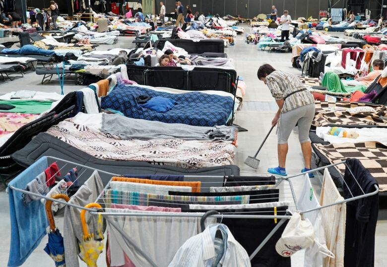 A woman sweeps the floor in a room filled with cots and people