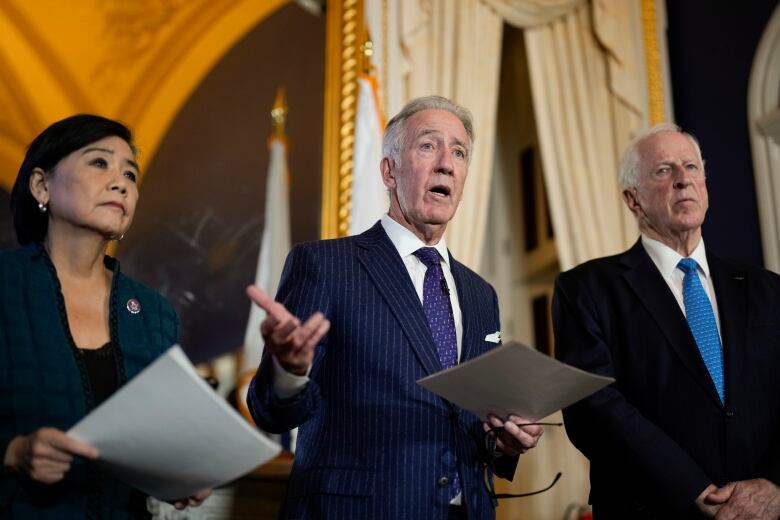 Three individuals wearing suits and holding documents speak to reporters. 