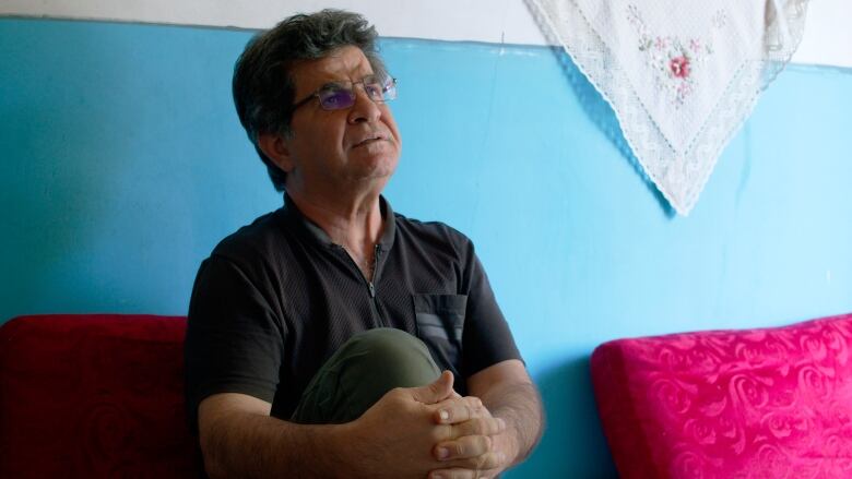 A man in a dark shirt sits, holding his bent leg, on a bright sofa against a blue wall.