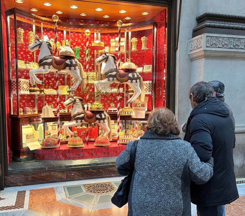 People stand on street outside of colourful window display showcasing panettone and carnival horses.