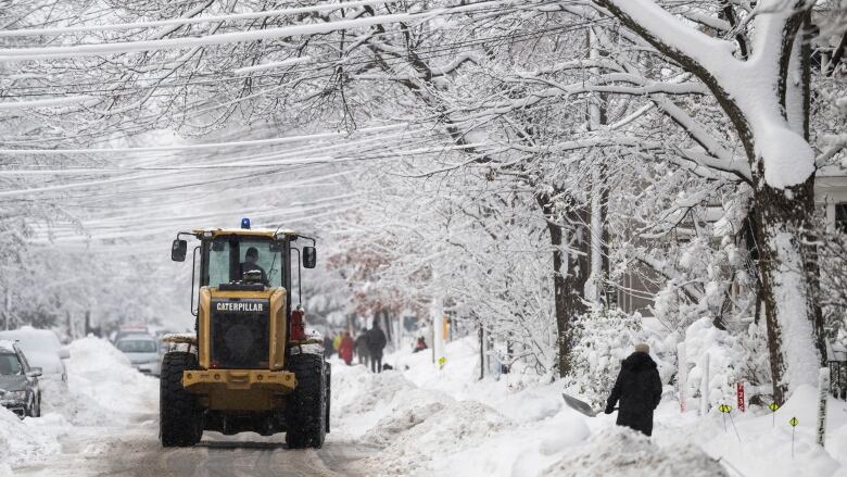 A snowplow is on the street, while someone is shoveling the sidewalk. There are large snowbanks on either side of the road, and trees covered in ice. 