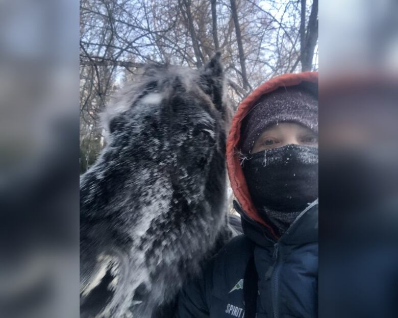 A woman with face cover and in jacket stands next to her horse, both covered with snow.