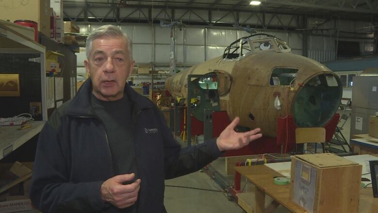 A grey-haired man wearing a black jacket stands in front of a non-functioning airplane in a museum warehouse. 