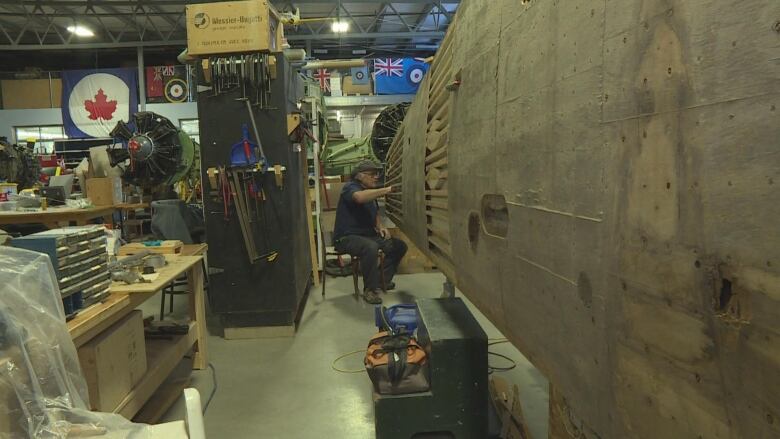 A white-haired man is seated next to a non-functioning airplane in a warehouse.