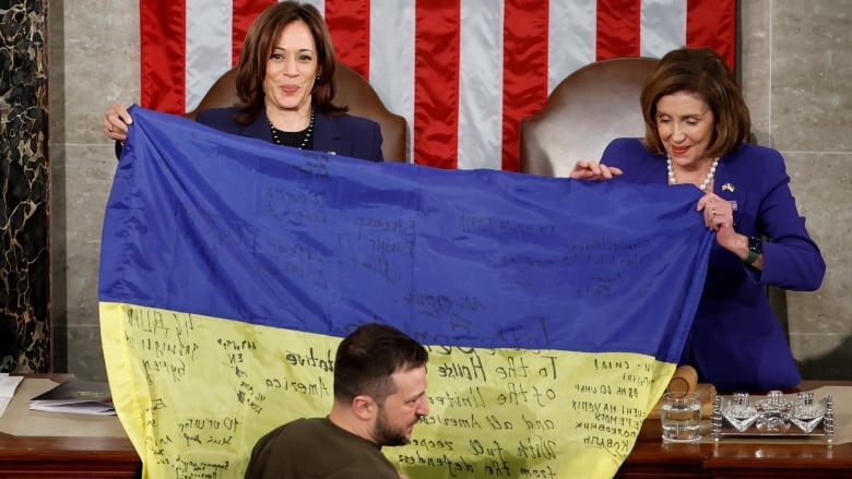 Individual in brown sweatshirt stands before a podium where two women hold a blue and yellow flag. 