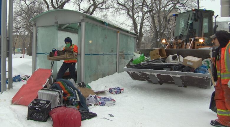 One man carries a box, while another man looks on with a shovel outside a bus shelter on a chilly day.