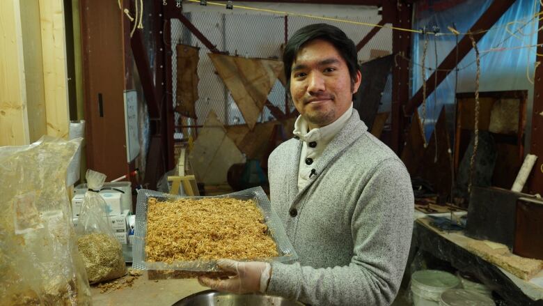 A male university student holds an oblong pan filled with mycelium and saw dust, which almost resembles a toasted coconut cake.