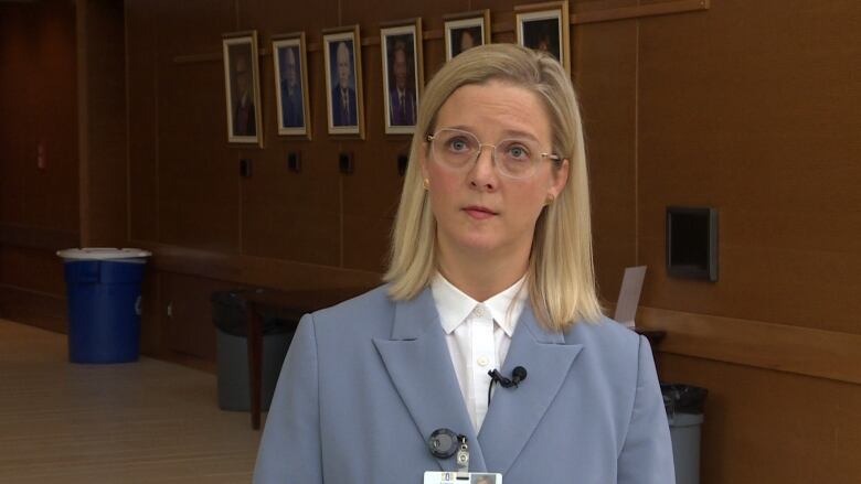 A blonde woman in a light blue blazer stands in front of the camera.