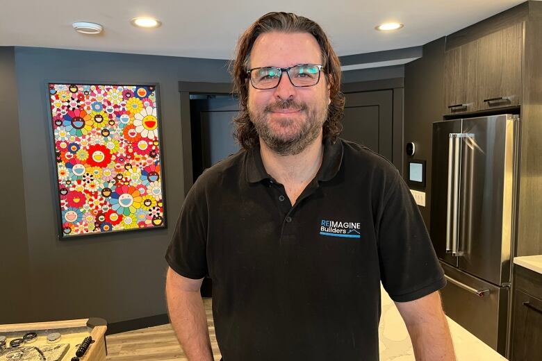 A man stands in his home, smiling, wearing a black polo t-shirt. There's a bright floral painting hanging in the background, on black walls.