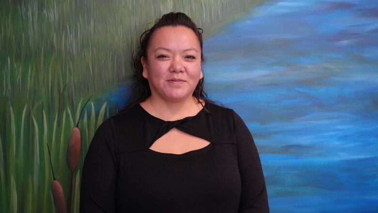 A smiling woman in black stands in front of a wall painting that features water and grasses.