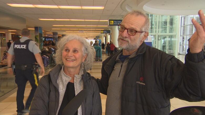 A woman with gray hair and a man with gray hair, a beard and glasses stand in an aiport.