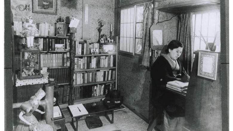 A woman sits writing a desk in a room with bookshelves.