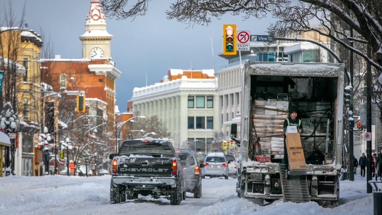 A person unloads a truck parked on the side of a snow-filled street. A lineup of cars waits, with a clock tower and snowy trees visible.