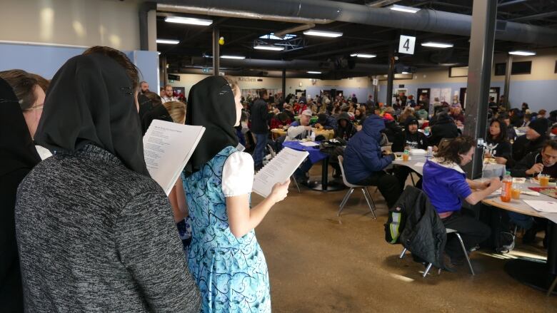 Women wearing colourful short-sleeved dresses and head coverings hold song books and sing while people eat at circular tables.