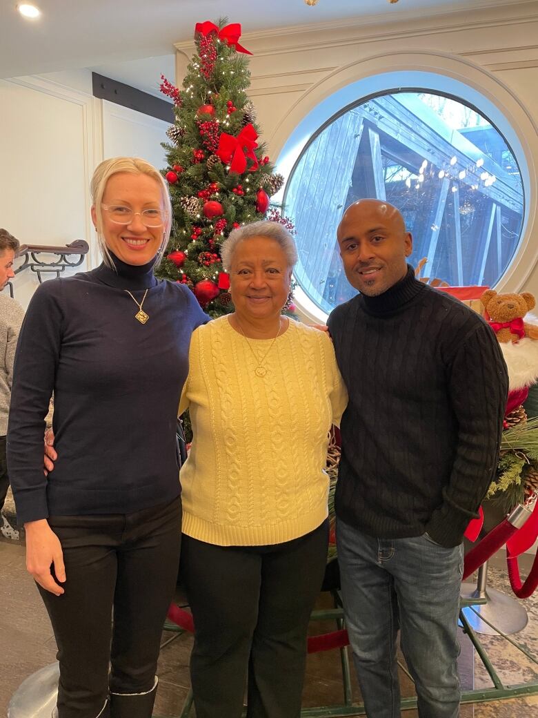 Three people smiling at the camera in front of a Christmas tree.