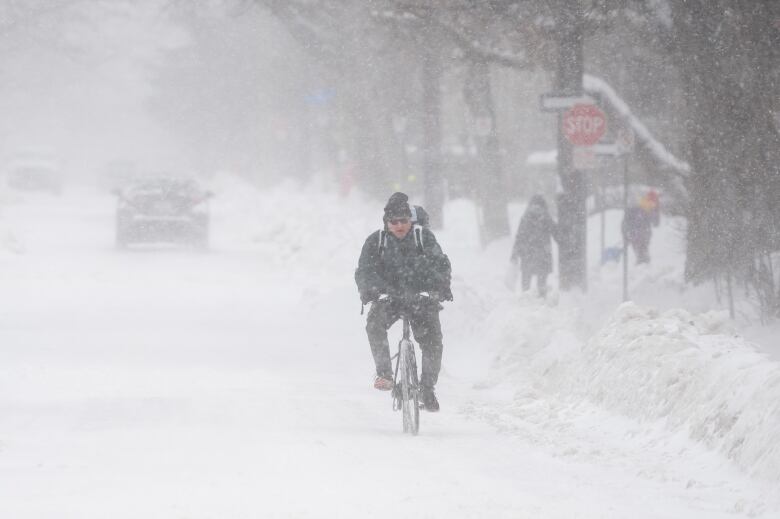 A man rides a bicycle down a street in the middle of a snowstorm.