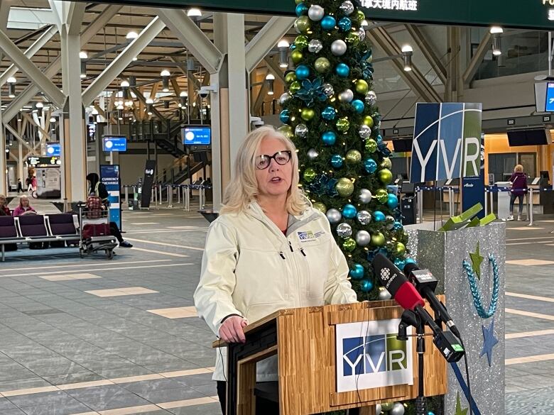 A white-haired woman stands at a podium at an airport, with a Christmas tree visible behind her.