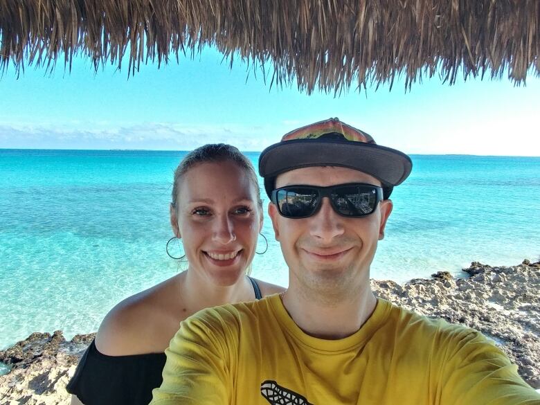 A man in sunglasses poses with a woman. Ocean can be seen in their background.