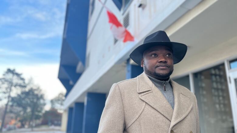 A Black man in a suit and dress hat stares to the right. Behind him is a frayed Canadian flag. 