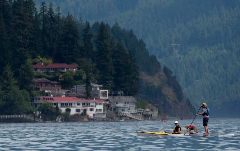 A woman and a dog on a stand-up paddle board are pictured next to another woman in a canoe off a coast, while cottages are seen on a hillside in the background.