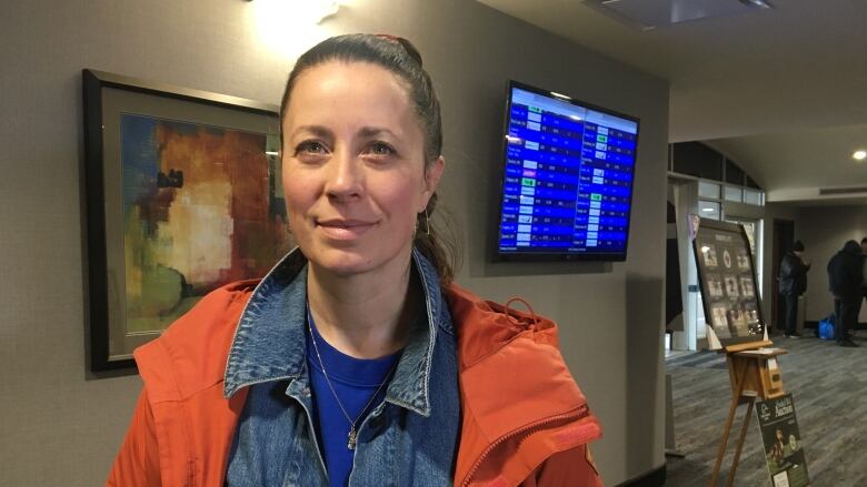 A woman with a ponytail in a blue sweatshirt and an orange jacket stands in front of a airport flight departure list.