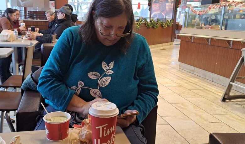 A woman sits in Tim Hortons with a coffee, soup and what appears to be a sandwich on the table in front of her.