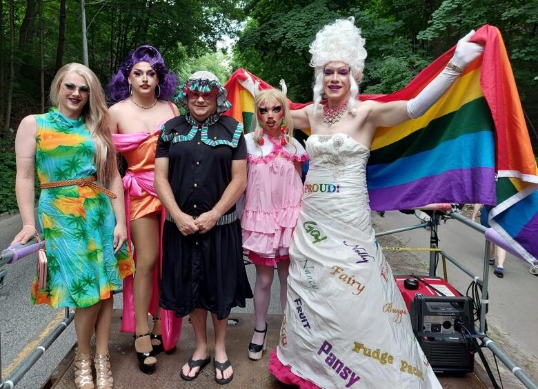 Five drag performers standing on a parade float with a rainbow flag draped around them.