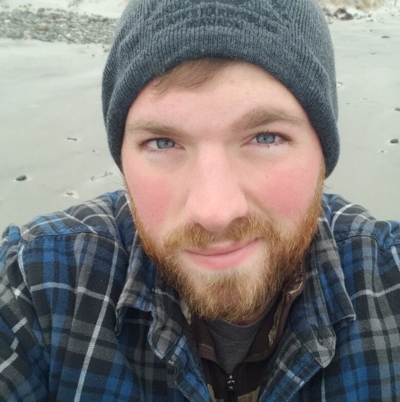 A man with a read beard is seen wearing a blue plaid button up and a dark grey touque.