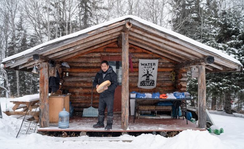 A man beats on a hand drum on the porch of a cabin in the winter.