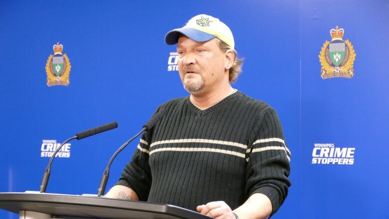 A man in a black sweater with a beige stripe wearing a ball cap stands at a podium with a Winnipeg police and Crime Stoppers banner in the background.