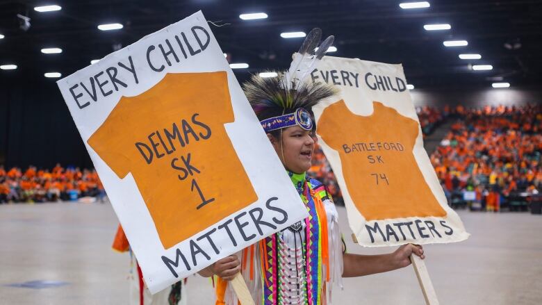 A man dressed in powwow regalia holds two giant Every Child Matters signs.