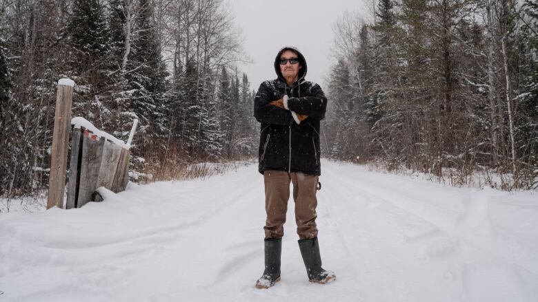 A man stands with his arms crossed in front of a snow covered road, with forest on either side.