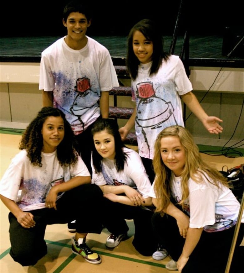 Four teen girls and one teenage boy wear matching T-shirts as they pose for a photo.