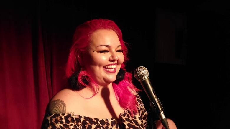 A woman with a large grin stands at a microphone in front of a red curtain.