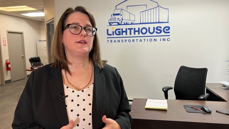 A woman is shown standing in front of an office desk with a sign on the wall that says Lighthouse Transportation which is the name of her company.