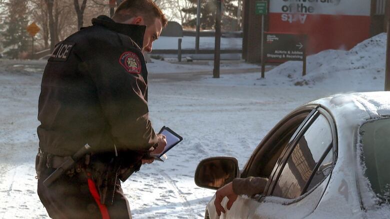 A uniformed Calgary Police officer leans over slightly to speak to a driver in a snow-covered sedan.