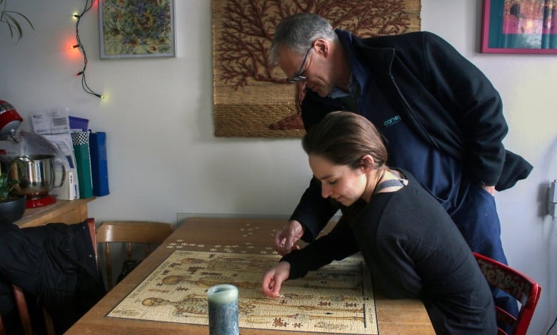 A young woman sits at a table doing a puzzle. Her dad stands behind her, helping her with the puzzle.