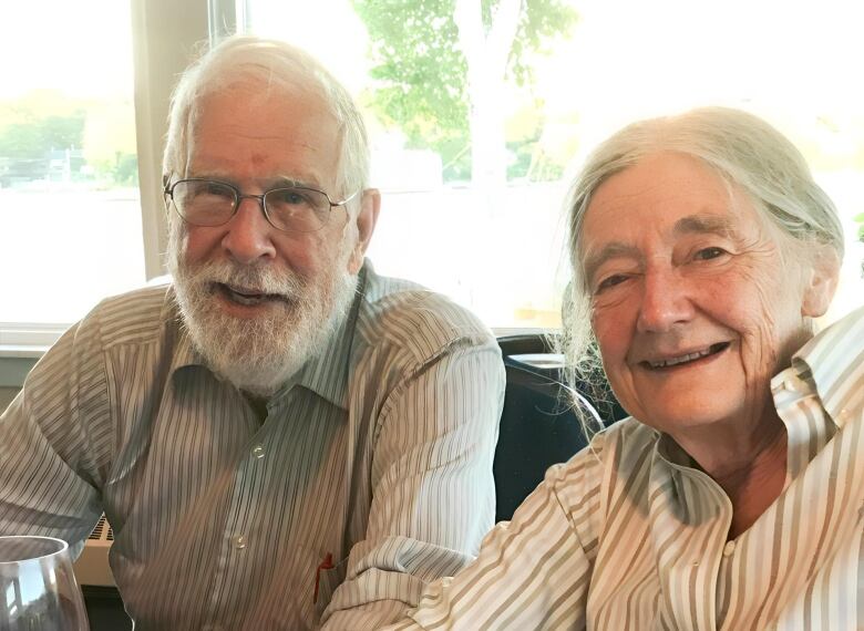 Alan Ruffman in a striped shirt on the right and his wife  Linda Christiansen-Ruffman, also in a striped shirt, smile at the camera against the background of a sunny window,