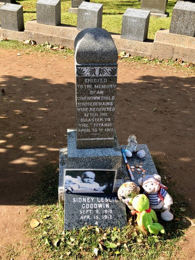 A grave marker in taupe granite memorializes the unknown child who died during the Titanic disaster in 1912. Below the marker is a plaque bearing the name Sidney Leslie Goodwin. There are a number of stuffed toys near the plaque.