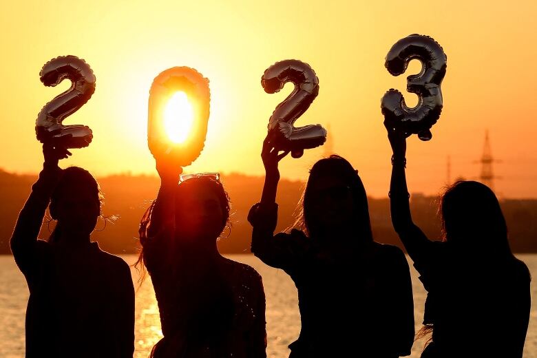 The women, facing the camera, hold up with their right hands individual balloons that together spell out '2023'. The golden sun is shining through the zero.