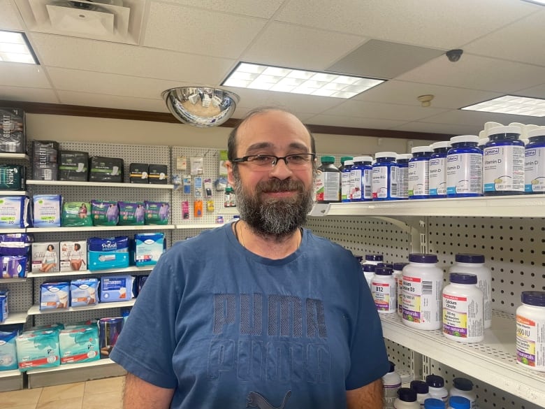 a man wearing glasses and a blue shirt smiling at the camera, in the background is a shelf of medicines at a pharmacy