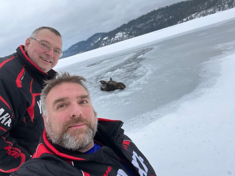 Two men take a selfie with a moose in the background. Only the moose's head is visible above a frozen lake.