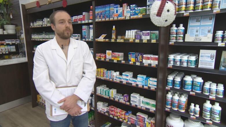 A man in a white lab coat inside a pharmacy holds a clipboard.